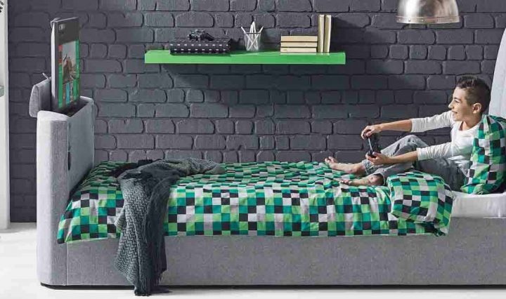 A teen using his bed with hydraulic TV lift to play video games.