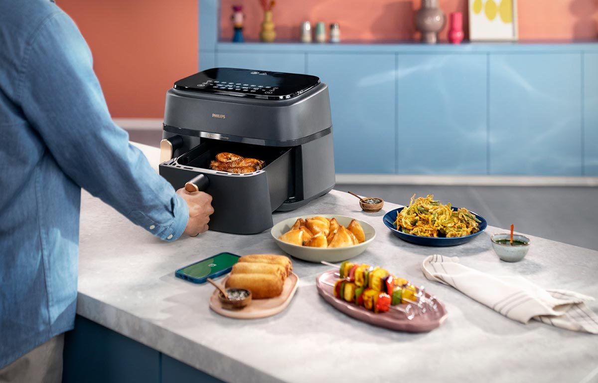A person opening the basket of a Philips 3000 Series air fryer, with an assortment of dishes laid out on a counter top to their right.