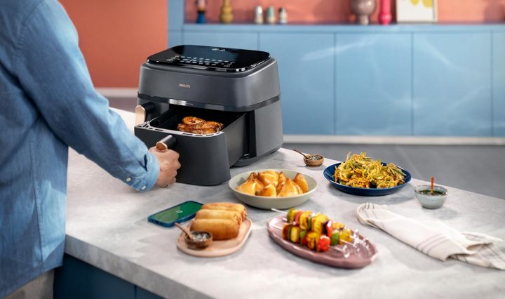 A person opening the basket of a Philips 3000 Series air fryer, with an assortment of dishes laid out on a counter top to their right.