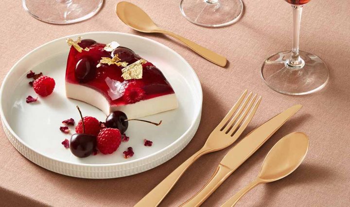 A plate with a slice of a jelly desert. The desert is two layered with the base layer being an opaque vanilla cream while the top layer is a red translucent jelly with raspberries inside. The desert is garnished with gold leaf and cherries.
