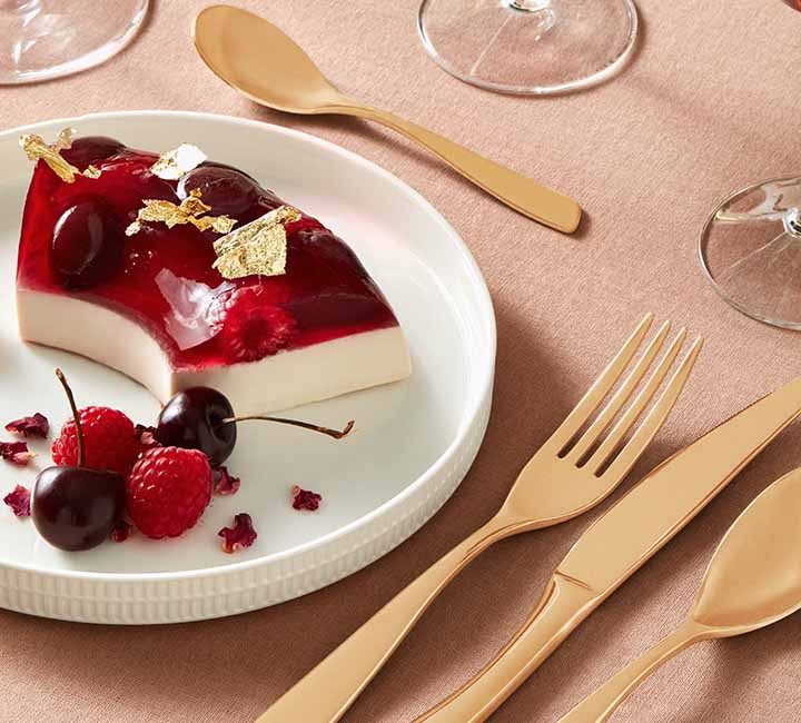 A plate with a slice of a jelly desert. The desert is two layered with the base layer being an opaque vanilla cream while the top layer is a red translucent jelly with raspberries inside. The desert is garnished with gold leaf and cherries.