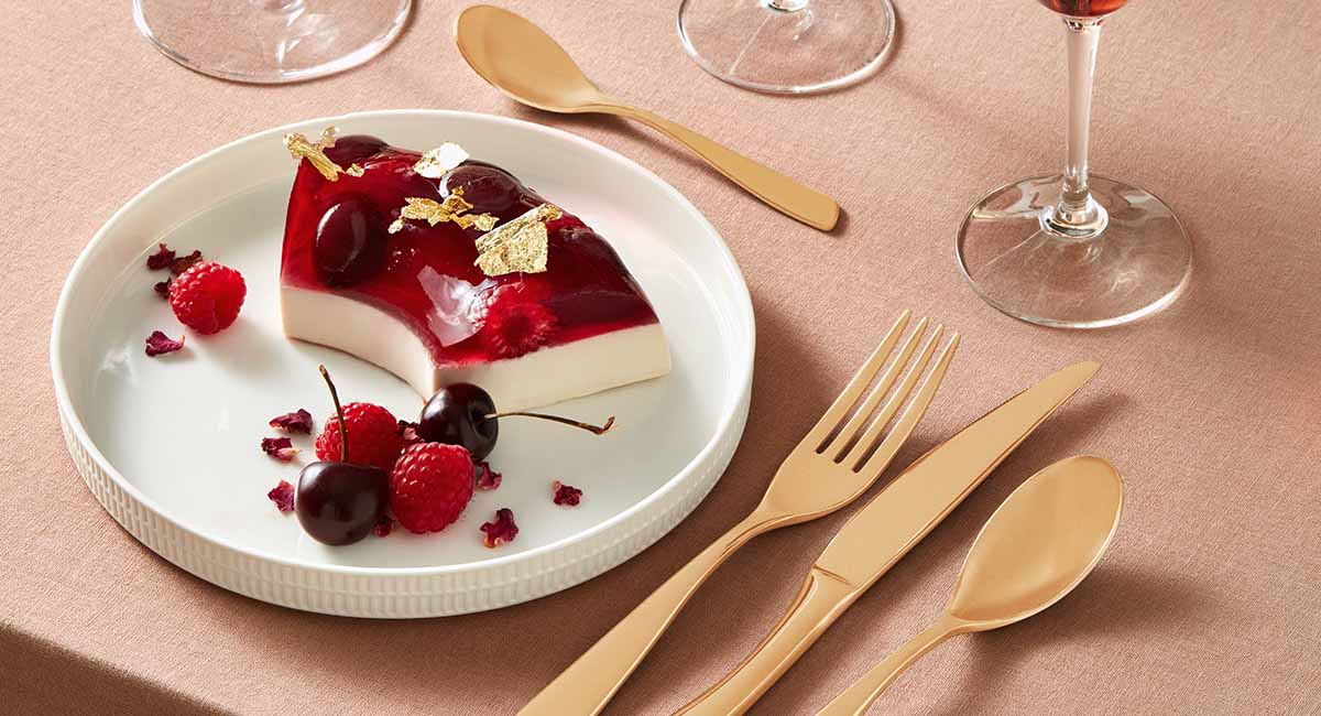 A plate with a slice of a jelly desert. The desert is two layered with the base layer being an opaque vanilla cream while the top layer is a red translucent jelly with raspberries inside. The desert is garnished with gold leaf and cherries.