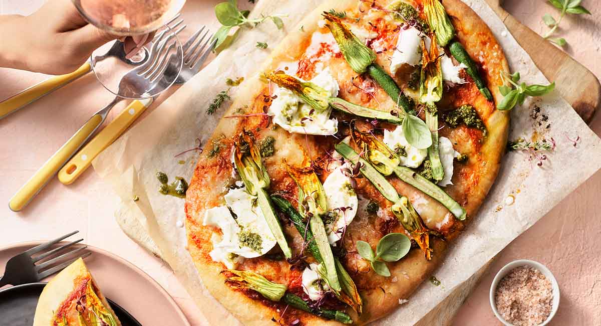 A pizza on a serving board. The pizza is topped with zucchini flowers, buffalo mozzarella and basil pesto.