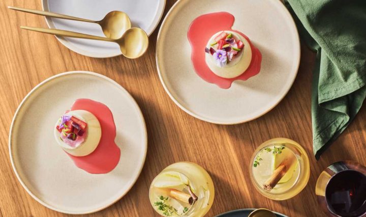 Overhead view of two bone-coloured plates set on a light wooden tabletop, each with a plated Jewelled Panna Cotta with Rose Petals & Pistachios.