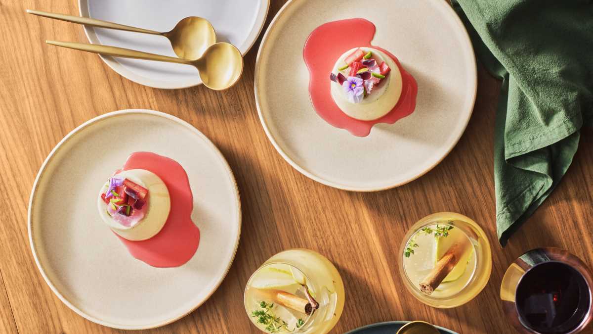 Overhead view of two bone-coloured plates set on a light wooden tabletop, each with a plated Jewelled Panna Cotta with Rose Petals & Pistachios.